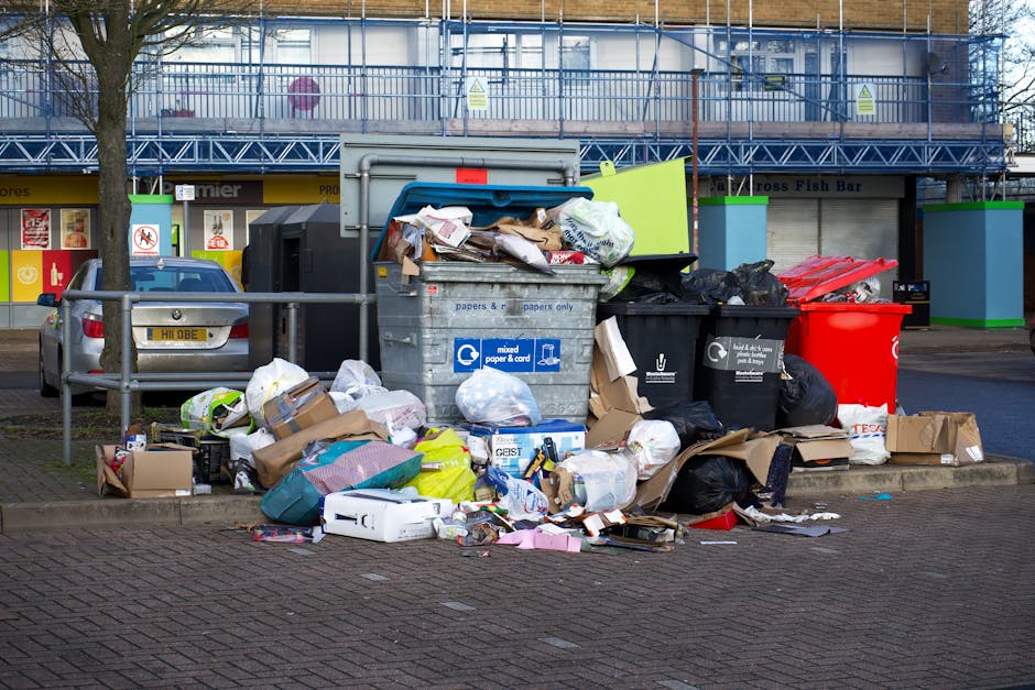 A collection of overflowing rubbish bins and assorted waste materials situated on a paved area beside a car and a metal railing, in front of a commercial building with blue scaffolding and signage. The waste includes a large grey mixed paper and card bin with its lid open, revealing crumpled paper, cardboard boxes, and plastic packaging. Next to it are black and red recycling bins filled with black plastic bags containing refuse. Cardboard boxes, some flattened and others partially assembled, are scattered across the ground, along with crumpled paper, plastic bags, and miscellaneous packaging items. A grey car with a visible UK registration plate is parked nearby, adjacent to the waste collection area. Behind the scene, the building features a shopfront with signs and a tiled facade, with a tree planted along the pavement. The environment appears to be a busy urban area, with natural daylight illuminating the scene and highlighting the cluttered state of the rubbish accumulation, which potentially relates to private waste disposal or alternative collection services often utilized in private house clearances or commercial waste management in the area.