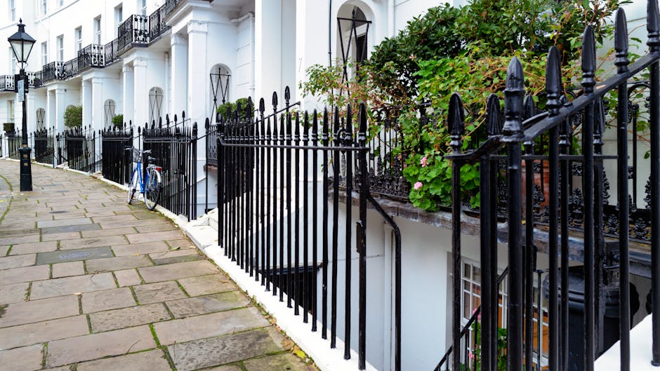 A row of white terraced houses with black wrought iron fencing along a paved sidewalk. The fencing features pointed finials and decorative scrollwork, separating the pedestrian pathway from the front gardens and staircases leading to the building entrances. A blue bicycle leans against one of the fences near the bottom left of the image. The front gardens contain lush green foliage and flowering plants, with some plants growing over the fence and spilling onto the walkway. The buildings have black-framed windows and narrow, curved staircases descending from the entrances, which are partly visible behind the fencing. Bright natural lighting illuminates the scene, highlighting the contrast between the white facades, black ironwork, and vibrant greenery. The image depicts a typical residential street with tidy, well-maintained pathways and the tidy organization of outdoor spaces, reflecting a setting suitable for private waste handling and on-site clearance activities as part of rubbish removal services offered by [COMPANY_NAME].