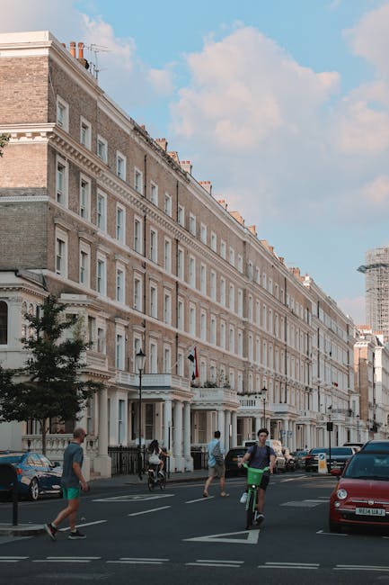 A row of four-storey Victorian-style terraced buildings with cream and light grey brick facades, decorative cornices, and sash windows, situated along a busy urban street. The buildings feature black wrought iron railings and white columns at the entrances, with small trees positioned in front of some properties. In the foreground, pedestrians are walking along the pavement, and a cyclist is riding on the street, which is lined with parked cars including a red vehicle on the right. The sky above is partly cloudy with patches of blue visible, and a construction scaffold is apparent on a building in the background to the right. The scene suggests a typical city residential area, where private waste or rubbish has been cleared, in the context of local rubbish removal services, such as those provided by House Clearances South Kensington. The environment appears clean, well-maintained, and active, with no visible rubbish or clutter in the street or on the sidewalks.