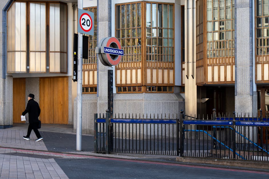 A narrow cobblestone street lined with mid-rise red brick buildings featuring white window frames and small balconies with wrought iron railings. On the left, there's a black sign with white text attached to a building, partially reading 'HEZZO', and a black street lamp with a decorative lantern installed on a pole. The street is bordered by short black bollards, and a few potted plants are visible along the sidewalk. The sky appears clear with natural daylight illuminating the area, creating subtle shadows on the buildings. The scene suggests an urban environment suitable for independent rubbish collection or private waste disposal, as no refuse or waste containers are visible but the setting reflects a typical location where rubbish removal services might be engaged to clear street clutter or prepare for waste collection in a residential or commercial district.