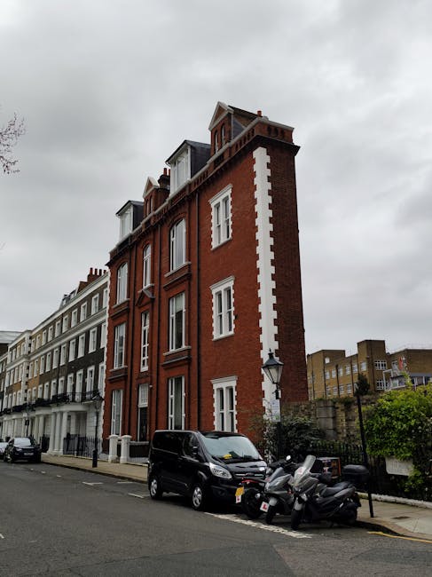 A multi-storey brick building located on a narrow street, featuring a mix of red and white brickwork with decorative white accents around the windows and along the corners. The building has a traditional architectural style, with small dormer windows on the top floor and a chimney visible against a cloudy sky. Parked directly in front of the building are several vehicles, including a black van and multiple scooters, which are situated on the street’s pavement. To the right of the vehicles, a small section of green shrubbery and a stone wall are visible, along with a street lamp mounted on a post. In the background, other residential buildings of varying heights and styles line the street, contributing to an urban residential setting. The scene suggests an area where private waste collection methods, such as house clearance services, may be employed rather than relying solely on local authority refuse disposal, aligning with independent waste handling solutions typical for urban terraces and narrow streets in South Kensington.