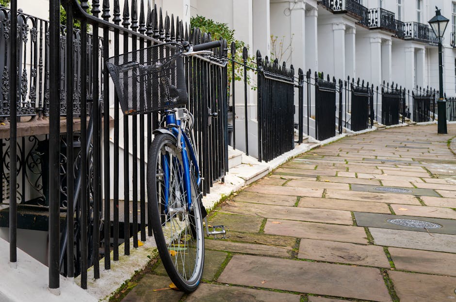 The image depicts a section of a residential street with a paved stone sidewalk running alongside a row of evenly spaced black wrought iron railings, which enclose front steps leading to white terraced houses with black wrought iron balconies. Leaning against one of the rails on the left side of the image is a blue bicycle with a black front basket, positioned upright with its kickstand on the uneven paved surface. The pavement appears weathered, with some moss or dirt visible between the stones. The background shows the facades of elegant townhouses featuring tall white columns supporting small balconies with decorative ironwork, and lantern-style streetlights mounted on the buildings. The scene suggests a typical urban setting where private waste collection or on-site clearance may be relevant, as the railings and pavement reflect an environment often associated with independent rubbish removal services. The lighting is natural, contributing to a clear view of the details, and the overall scene is tidy and orderly, with no visible waste or clutter in the surroundings.