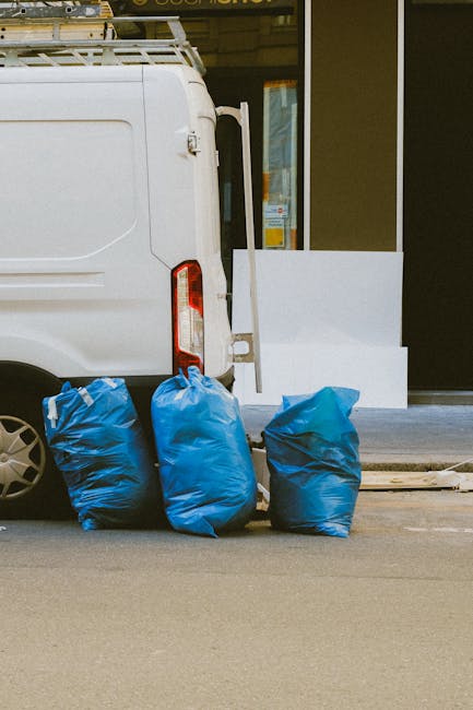 Three large blue rubbish bags filled with waste are lined up next to the rear of a white delivery van parked on a street, with the back doors closed and a small section of the vehicle's tail light visible. The bags are made of plastic with a crinkled texture and are positioned on the pavement, which shows minor scuff marks. Behind the van, there is a modern storefront with large glass windows and doors, surrounded by a dark frame, and a white rectangular object—possibly a piece of bedding, panel, or shelving—leans against the building wall on the right. The environment appears to be an urban area, likely near a commercial or residential premises, with the scene suggesting an instance of private rubbish disposal or clearance, possibly coordinated through a waste removal service such as House Clearances South Kensington, as indicated by the context. Lighting is natural, with soft shadows cast on the pavement, illustrating a clear day suited for outdoor waste collection activities.