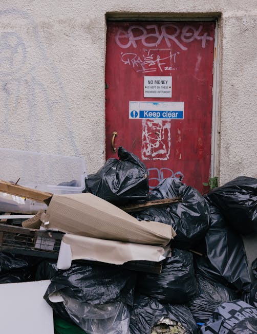 A collection of black rubbish bags, some partially torn, piled up in front of a red metal door set into a beige, textured exterior wall. The bags contain household waste and are stacked unevenly, with some leaning against the wall and others spilling onto the ground. Several pieces of discarded packaging and broken cardboard are visible on top of the pile. The red door features graffiti markings, a white sign with black text indicating 'NO MONEY' and 'KEEP CLEAR,' and a blue sticker with the phrase 'Keep clear.' The environment suggests an urban alleyway or service area where waste is temporarily stored before collection, reflecting a scene of private waste disposal outside a property, possibly managed by a waste removal service such as House Clearances South Kensington. Subtle signs of grime and weathering on the wall and door highlight the practicality of on-site rubbish storage before mainstream collection or disposal.