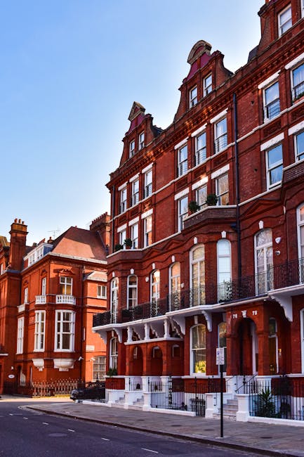 A row of Victorian-style red brick residential buildings along a city street, featuring large sash windows with white frames, ornate wrought iron balconies on the first-floor windows, and decorative stone cornices. The buildings have prominent pitched roofs with dormer windows, and some include small front gardens enclosed by low metal railings. The pavement in front is clean and free of obstructions, while a black litter bin is visible near the curb. The scene is illuminated by natural daylight, with a clear blue sky overhead, and the overall environment suggests an urban residential area where independent rubbish collection or on-site clearance might be relevant to manage waste in such property types, relating to private disposal services like those provided by houseclearancesouthkensington.co.uk.