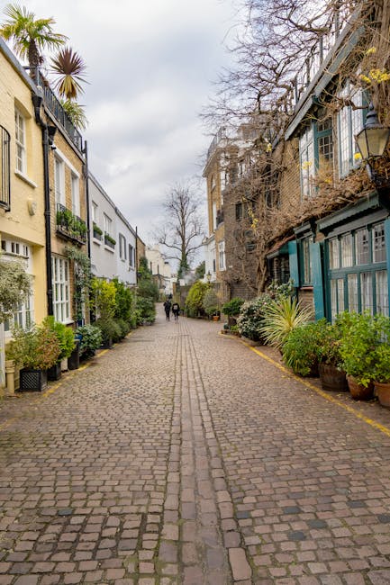 A narrow cobblestone street in a residential area of South Kensington, featuring a mix of traditional terraced houses and newer buildings on either side. The left side showcases cream-colored facades with large windows, some with flower boxes, and a tall palm tree visible behind the buildings. The right side displays a weathered brick building with blue-painted wooden balconies and window frames, partially covered with climbing plants and leafless branches. Potted shrubs and plants line both sides of the street, creating a green border. The street appears to be in a quiet, historic neighbourhood with a few pedestrians walking in the distance under an overcast sky, evoking a typical scene where private waste disposal or independent rubbish removal services like those offered by houseclearancesouthkensington.co.uk might be relevant for maintaining such urban environments.
