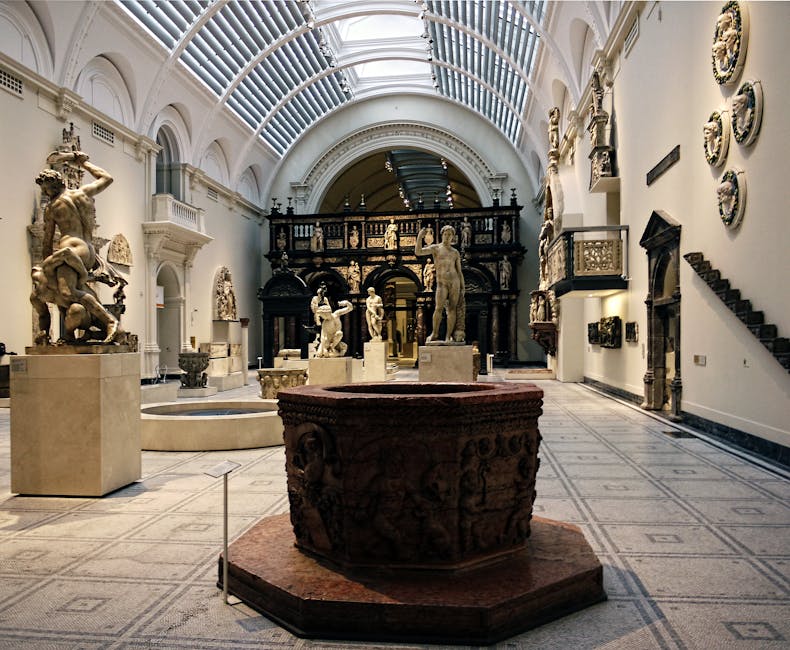 An interior view of a museum gallery with a high, arched glass ceiling allowing natural light to illuminate the space. The room features classical sculptures made of white marble, displayed on raised pedestals, including a group of figures and individual statues, some with detailed drapery and anatomical features. The sculptures vary in size and are arranged across the tiled floor, with a larger, dark-colored carved stone basin or decorative object in the foreground positioned centrally. The walls are decorated with circular plaques or reliefs, and a black ornate balcony with intricate railings extends along one side. In the background, an elaborate dark wood or metal architectural structure frames an arched doorway, leading to another section of the museum. The clean and well-maintained environment reflects a typical setting for cultural and historical exhibitions, with no visible rubbish or waste present, consistent with professional museum care and maintenance, as would be handled by services such as houseclearancesouthkensington.co.uk specializing in interior space waste management and collection.