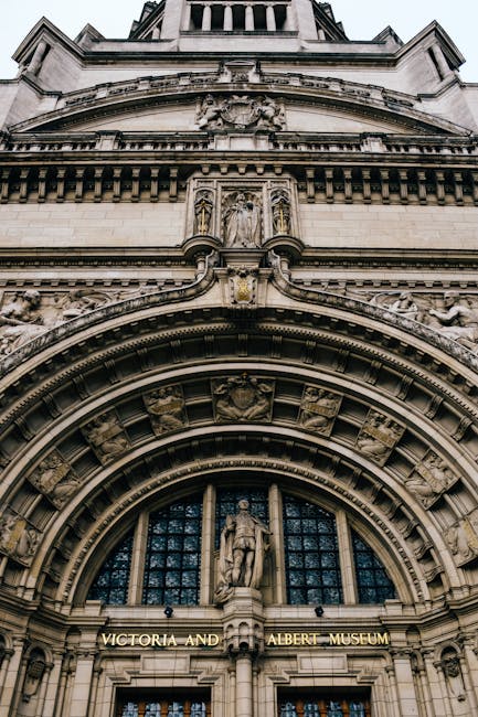 A close-up view of the ornate stone facade of the Victoria and Albert Museum in South Kensington, showing intricate architectural details including carved sculptures and decorative arches above the entrance. The image captures the building's historic stonework with a focus on the detailed carvings and religious figures positioned above the doorway, illuminated by natural daylight that highlights the textures and finishes of the aged stone. The perspective emphasizes the grandeur of the structure, with a glimpse of the upper levels extending out of view, set against a clear sky. The sophisticated craftsmanship and historic character of the building reflect its cultural significance, and the context of independent or private waste removal services, such as those provided by houseclearancesouthkensington.co.uk, are relevant for aesthetic and logistical considerations when managing waste in heritage sites or similar urban environments.